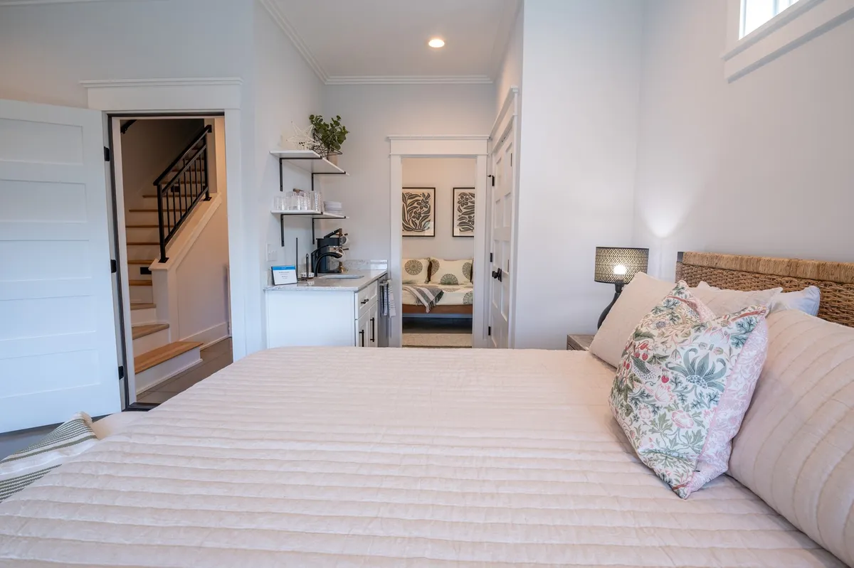 This scene shows a cozy bedroom with a bed adorned with decorative pillows, a convenient kitchenette area, a staircase leading to another floor, and modern artwork on the wall. The soft light filtering through windows enhances the welcoming atmosphere.