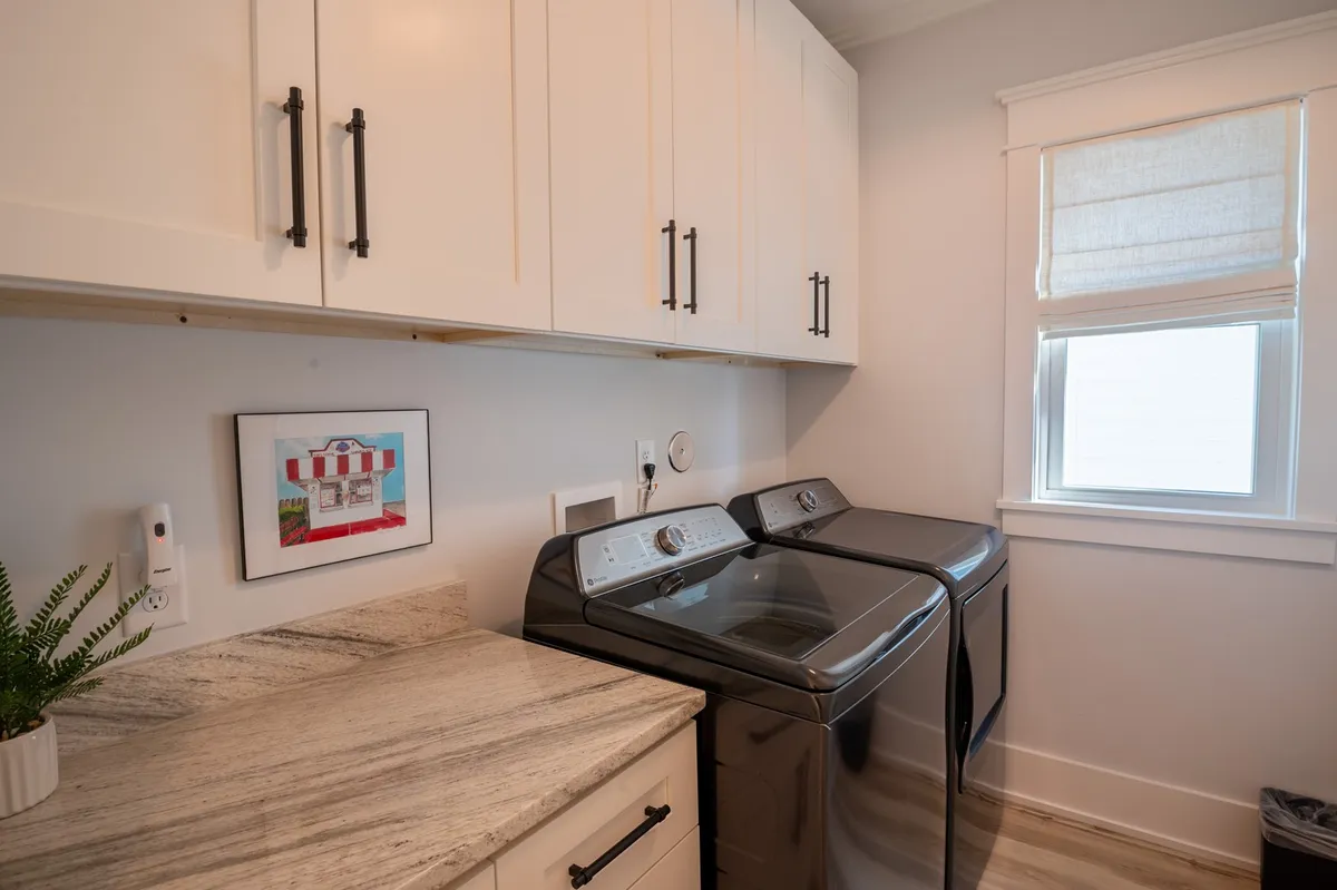 A bright and practical laundry room is shown with a high-capacity washer and dryer, a granite folding counter, framed artwork, and a large window letting in natural light.