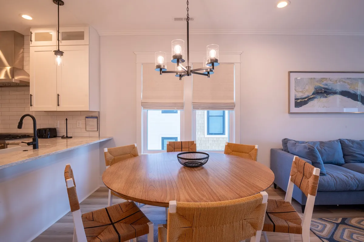 A round wooden dining table set under an elegant chandelier, with a comfortable sofa and tasteful wall art in the background. The area feels open, inviting, and ideal for both meals and casual conversation.