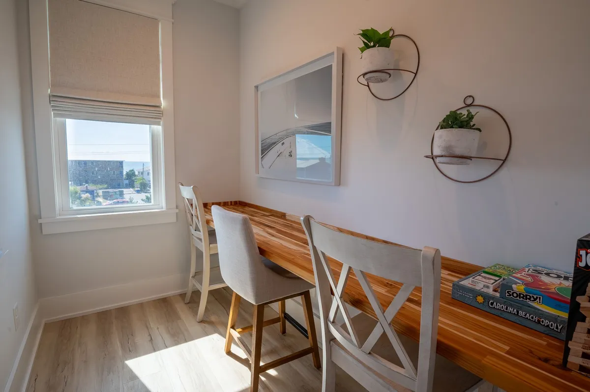 A sunlit office nook with a wooden table, potted plants, a window view, and board games, creating a relaxed atmosphere for work or leisure. The space blends productivity and comfort with a touch of playfulness.