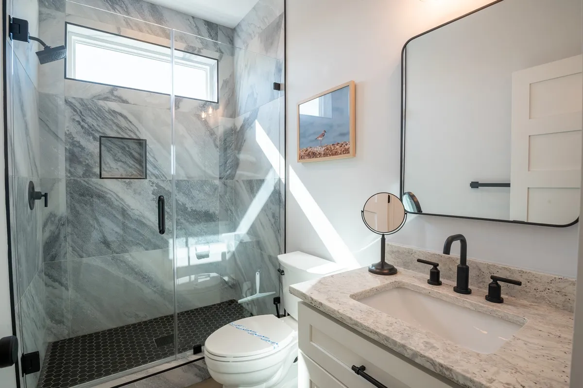 A bright, fresh bathroom highlights a glass shower enclosure, marble wall tiles, a granite countertop, black fixtures, and framed artwork. The design exudes spa-like luxury, ideal for self-care routines and relaxation.