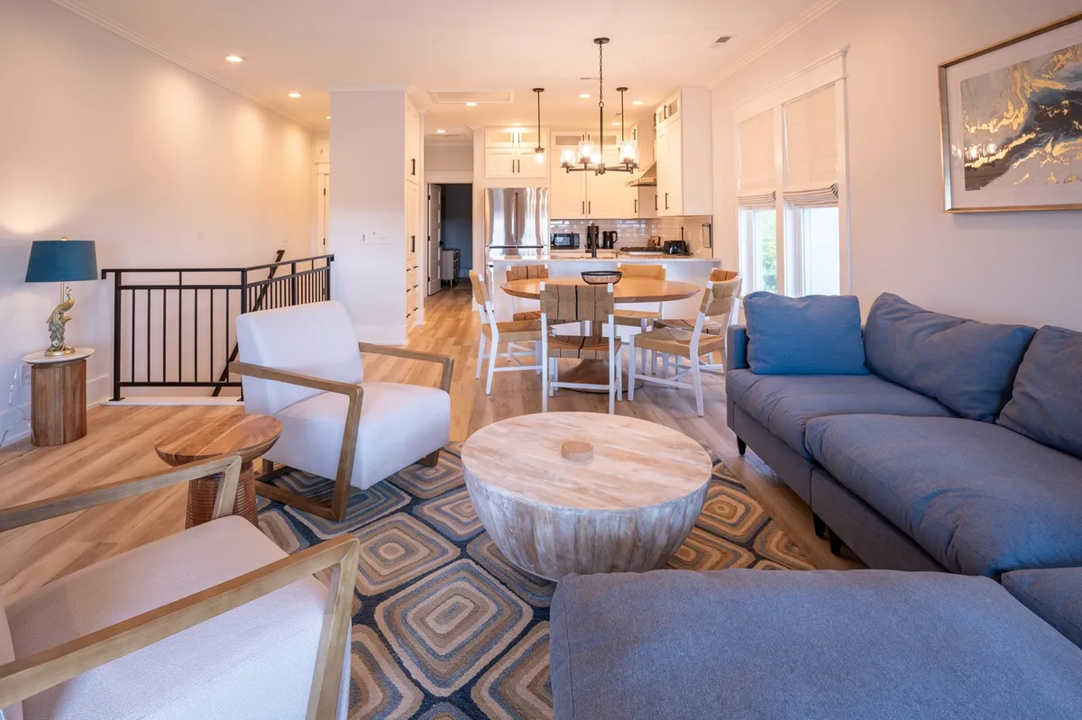 The image showcases a sunlit, open-concept living and dining area with a plush sectional sofa arranged around a round wooden coffee table, and a spacious dining table with enough seating for family or group gatherings. Natural wood accents and an abundance of natural light create a warm, inviting atmosphere ideal for relaxation and togetherness.