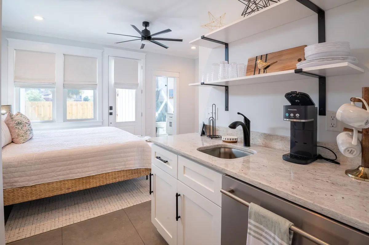 A cozy bedroom corner highlighting a bed, a coffee maker on open shelving, and plenty of natural light from a nearby window. The space feels welcoming and perfect for starting the day with a fresh brew.