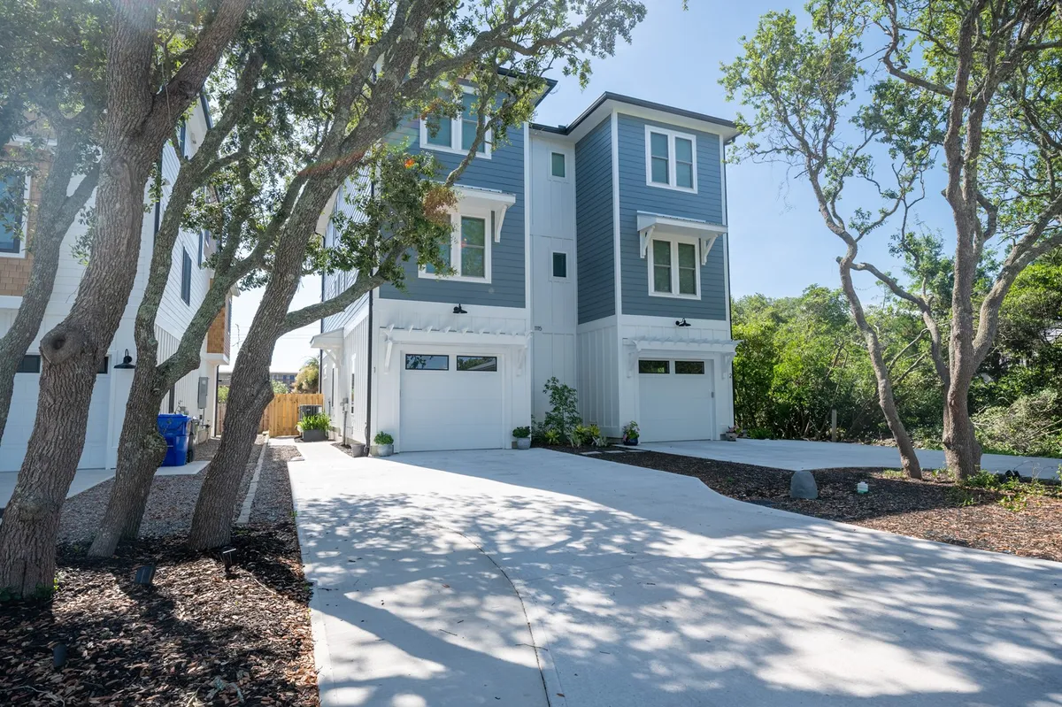 A sunlit, two-story house framed by mature trees, with a welcoming driveway and expansive porch areas ideal for gathering. The vibrant setting hints at the proximity to the beach and offers a glimpse of the relaxed outdoor lifestyle perfect for families.