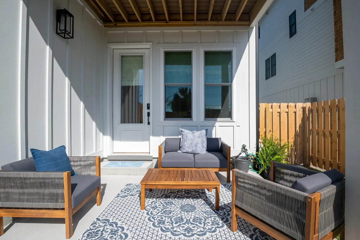 A cozy, sun-drenched exterior seating area featuring a decorative rug, wooden coffee table, and potted plants. This space is designed to foster relaxation and casual socializing, with ample seating that invites guests to linger and enjoy the outdoors.