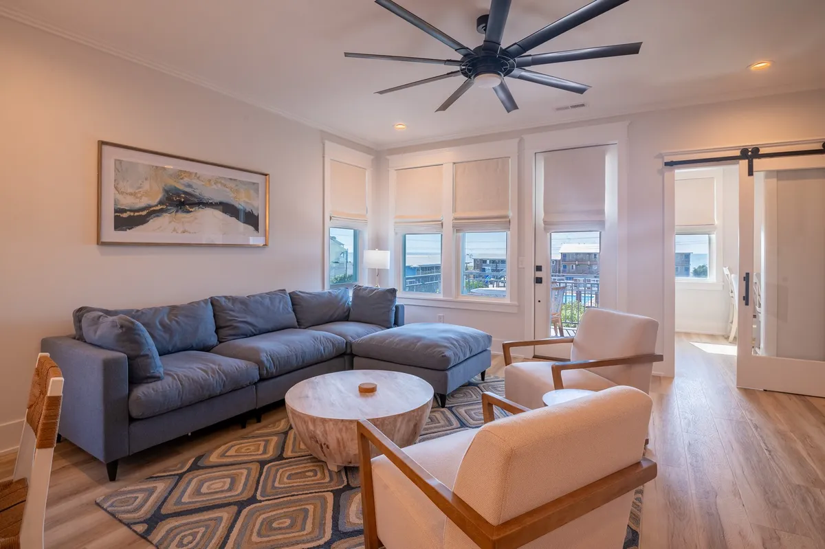 A spacious living area with a cozy sectional sofa, round coffee table, accent chairs, and large windows that flood the room with natural light. The arrangement invites groups to relax, converse, or play games, with sunset views hinting at a seamless connection to the outdoors.