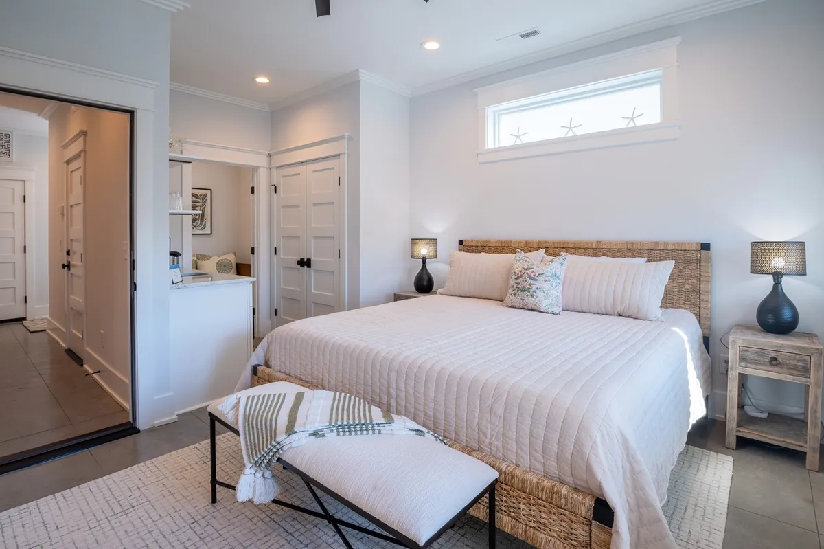 This image features a king-sized bed with a woven headboard, nightstands with lamps, decorative pillows, and a bench at the foot of the bed. The mood is serene and restful, with soft natural light illuminating the space, creating a sense of luxury and tranquility.