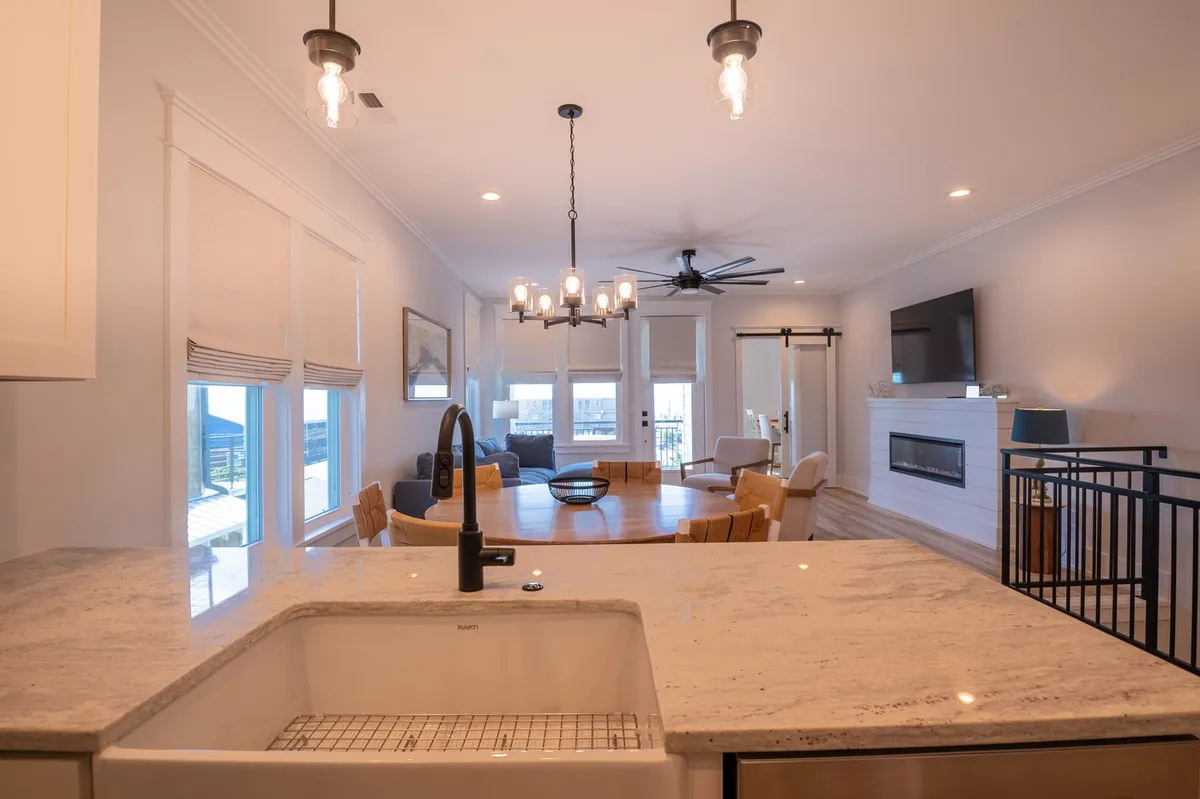 A spacious open-plan great room with a large kitchen island, a welcoming dining table, a stylish chandelier, and a cozy fireplace. The space is bright and airy, ideal for gatherings, with premium furnishings and a warm, communal atmosphere.