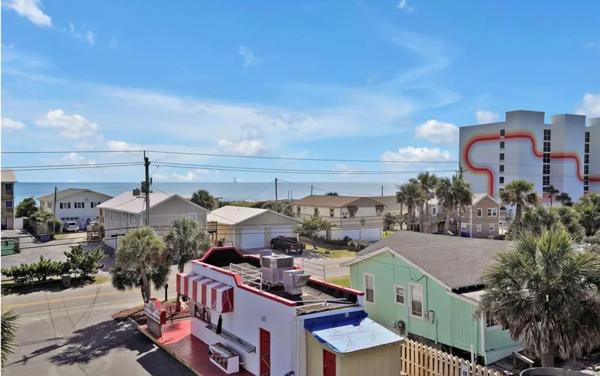 A vibrant street scene with colorful buildings, a sparkling ocean view, and lush palm trees. People are seen strolling, exploring local shops, or heading towards the beach, capturing the lively atmosphere of Carolina Beach's boardwalk and attractions.